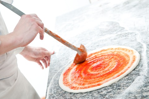 Pizzabodem op werkblad terwijl een kok tomatensaus gelijkmatig verspreidt met een lepel