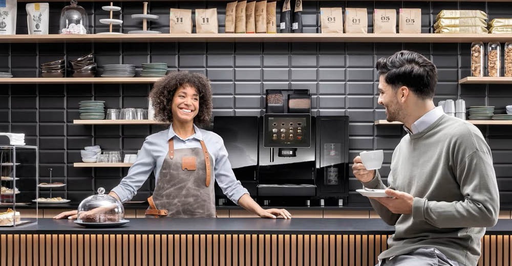 Vrouwelijke barista achter de toonbank van een moderne koffiebar glimlacht naar een klant die op een kruk zit met een kop koffie in zijn hand.